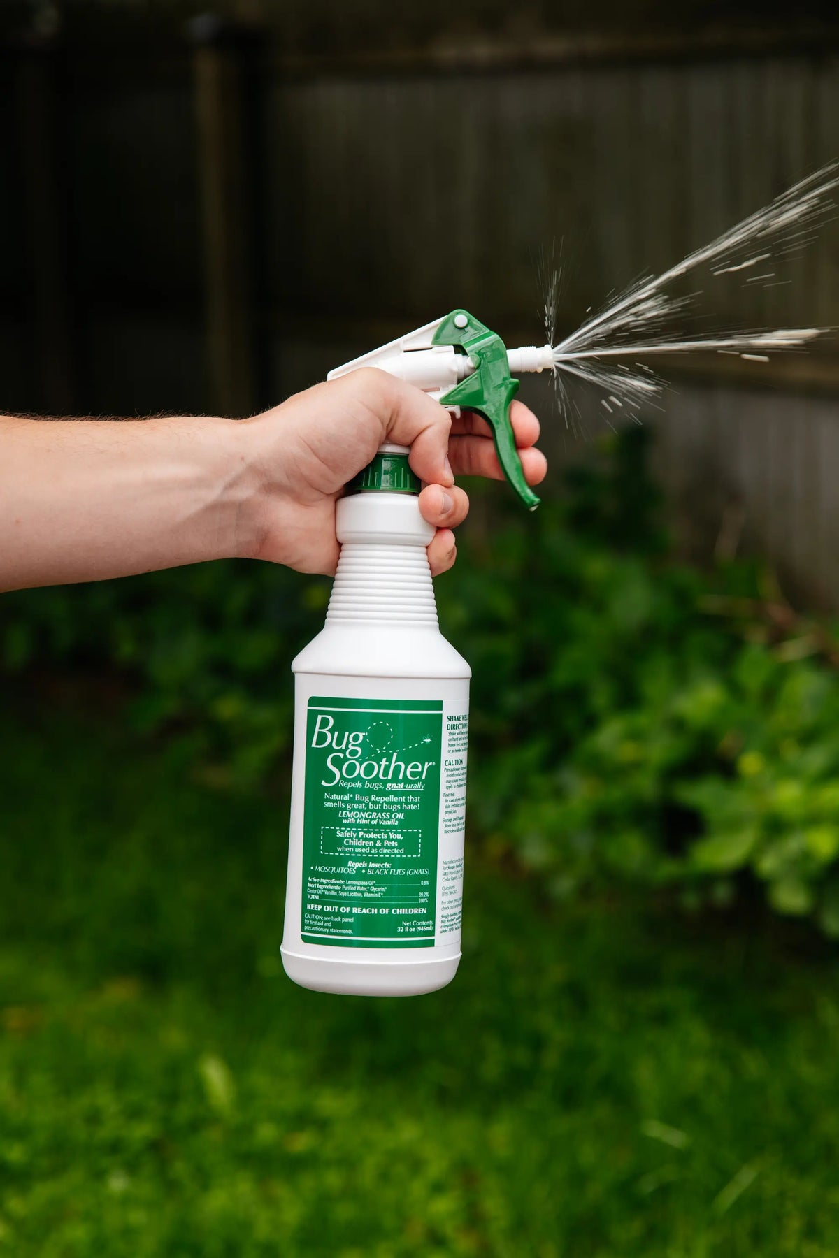 A hand holding a white spray bottle labeled 'Bug Soother' with a green trigger sprayer, spraying a fine mist outdoors in a grassy garden area.