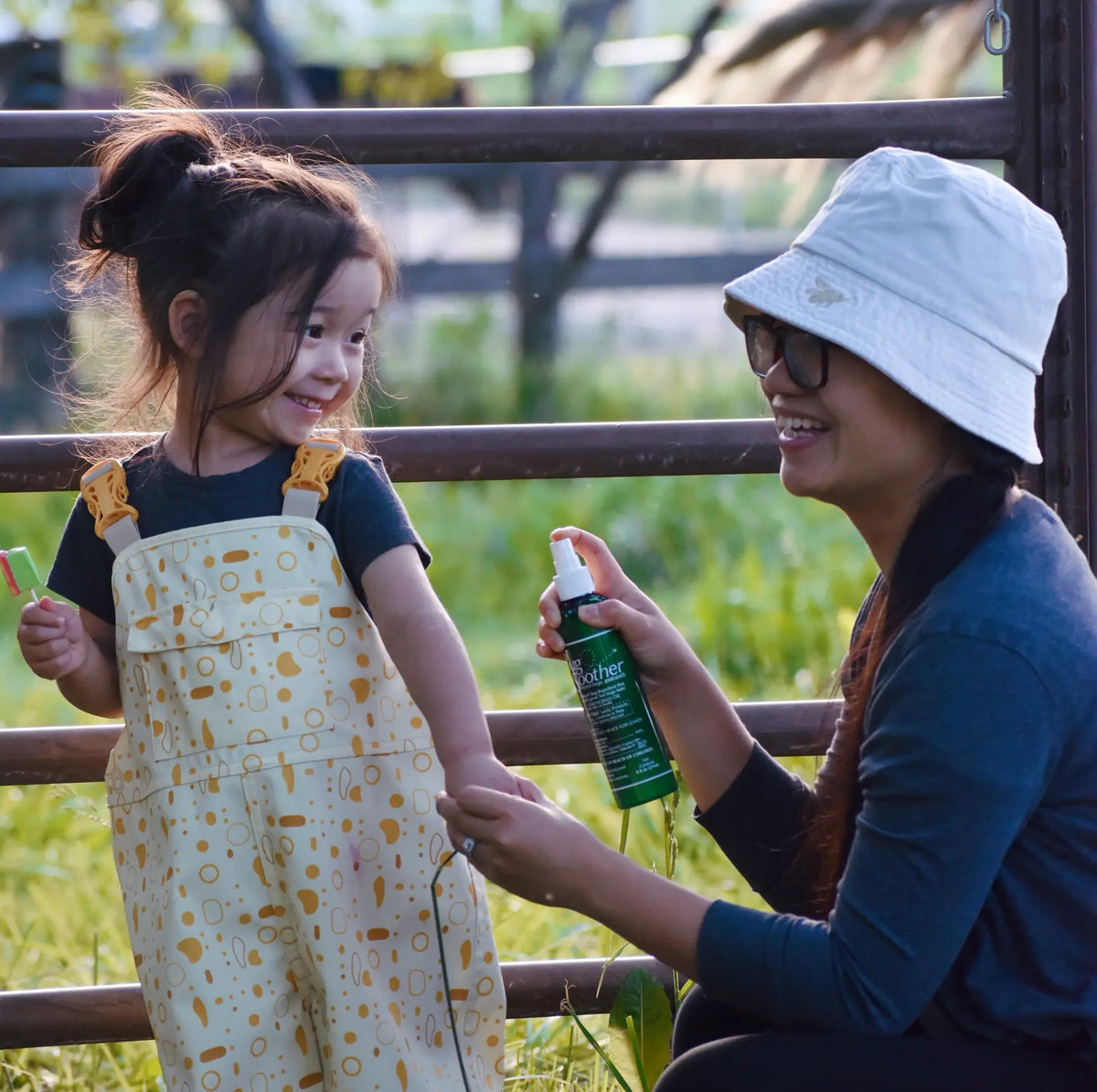 A woman wearing a white bucket hat and sunglasses sprays a child wearing a cream and gold patterned bib overalls outdoors next to a wooden fence.