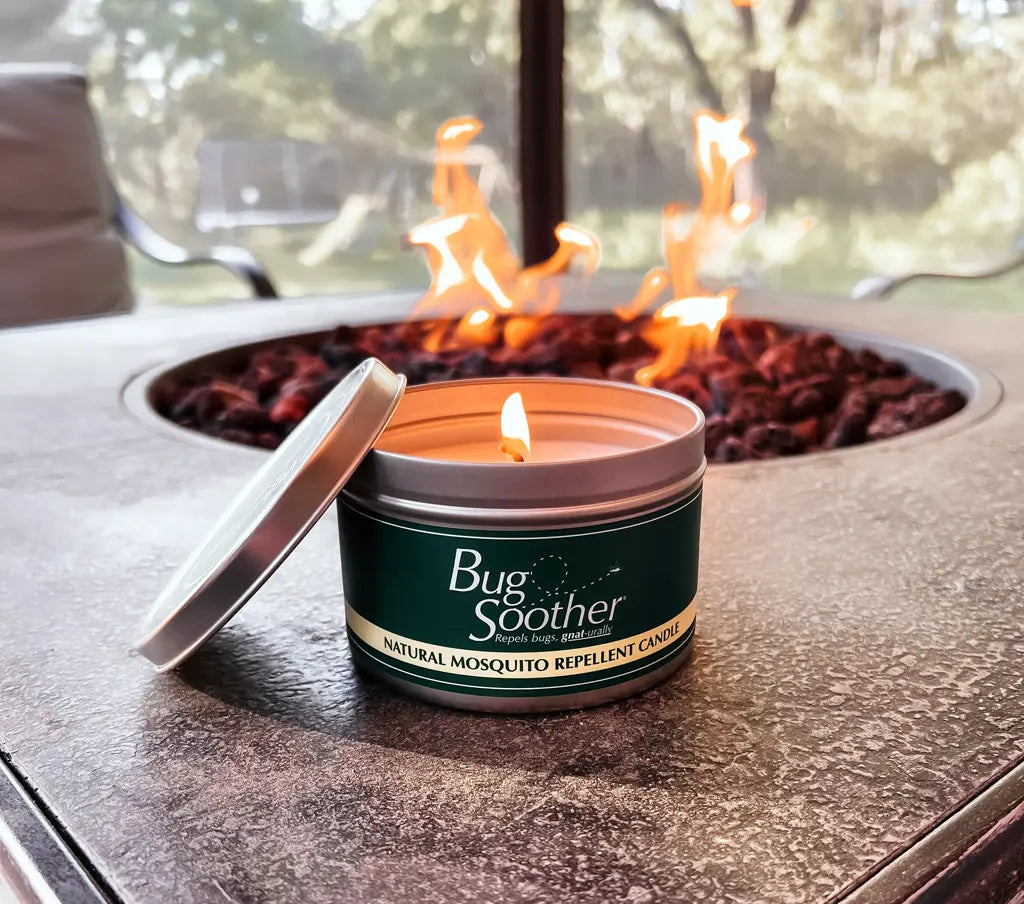 A lit Bug Soother candle on a outdoors table with a firepit 
