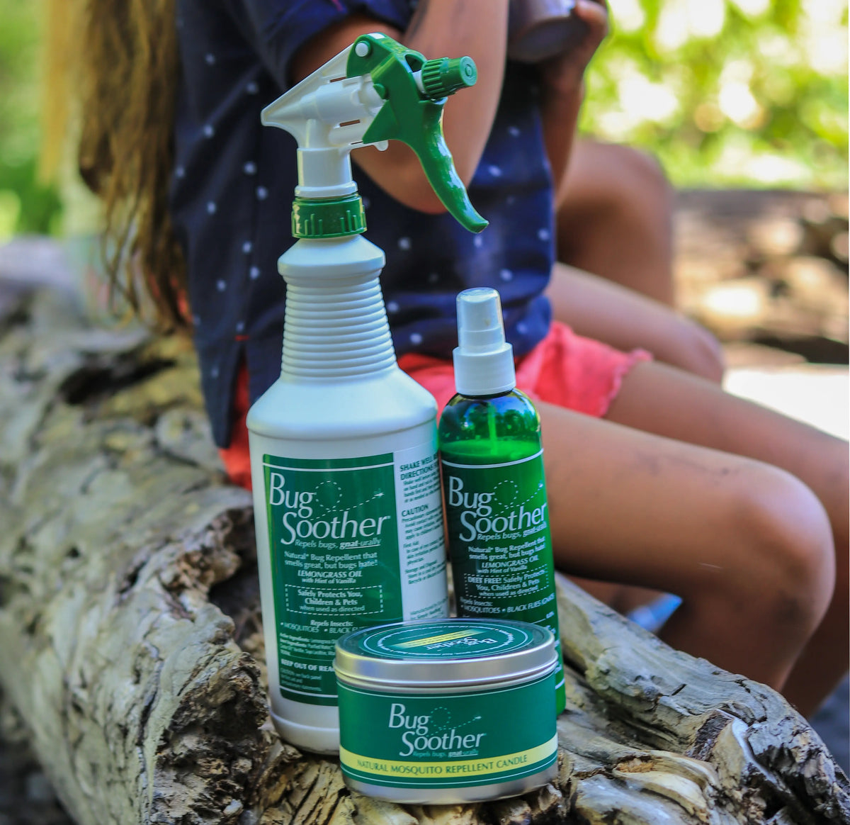 Three Bug Soother products arranged on a log outdoors: a large white spray bottle with a green sprayer, a green spray bottle, and a round tin with a green lid. A child is seated in the blurred background.
