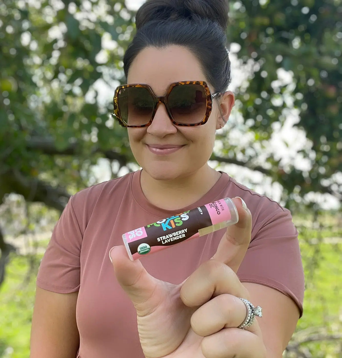 A woman holding up an Mongo Kiss Strawberry Lavender lip balm. 